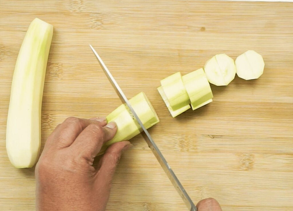 Two peeled courgettes on a wooden chopping board. One courgette is whole, the other is being sliced by a woman