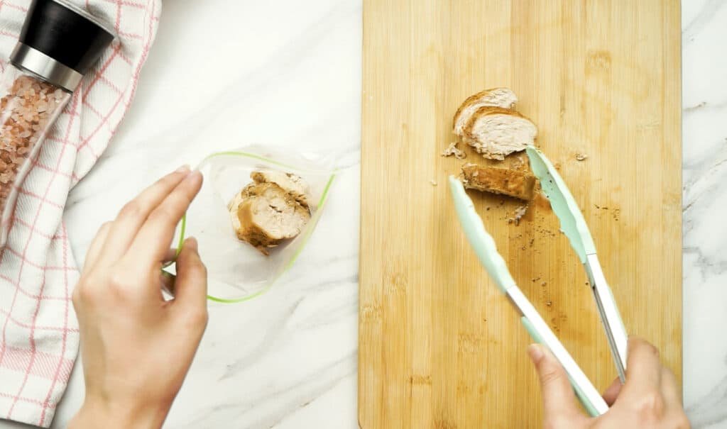 A woman's hand holding a freezer bag open containing slices of chicken breast as uses green tongs to add more slices to the bag