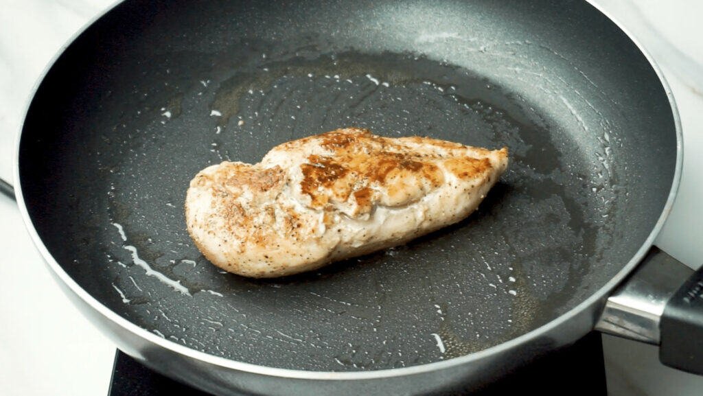 A chicken breast being cooked in a non-stick pan with a little olive oil