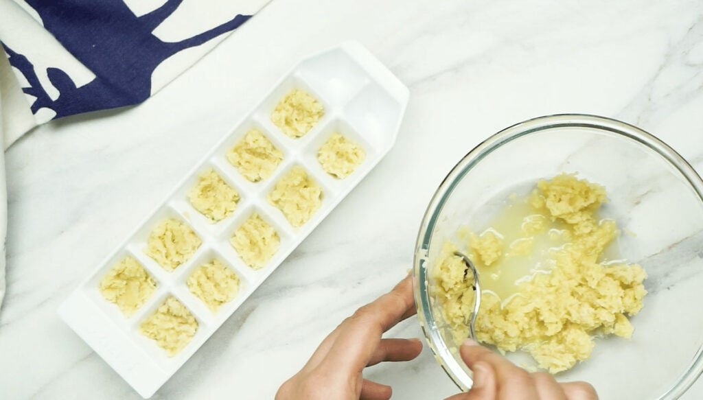 Grated ginger in a glass bowl being scooped up with a dessert spoon to be added to a white ice cube tray