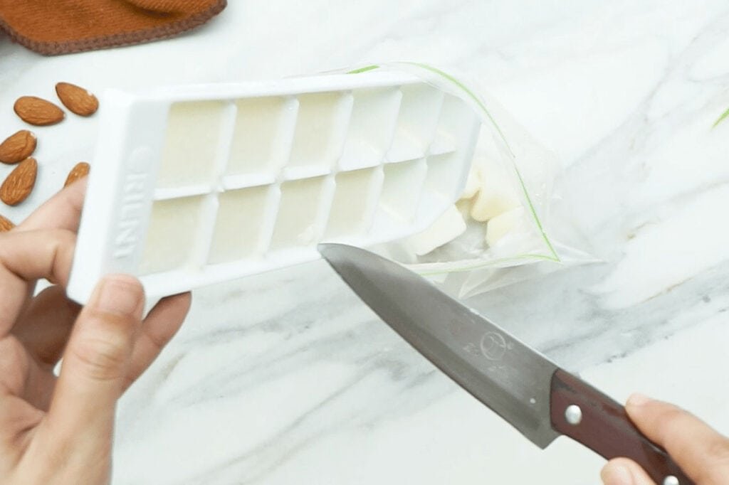 Frozen cubes of almond milk being removed from an ice cube tray and tipped into a freezer bag