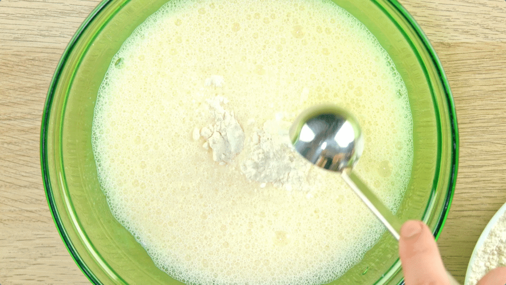 Flour being added to whisked eggs and milk in a green-tinted glass bowl