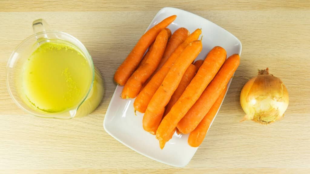 3 ingredients required to make carrot soup showing vegetable stock in a glass jar, carrots on a white plate and a yellow onion