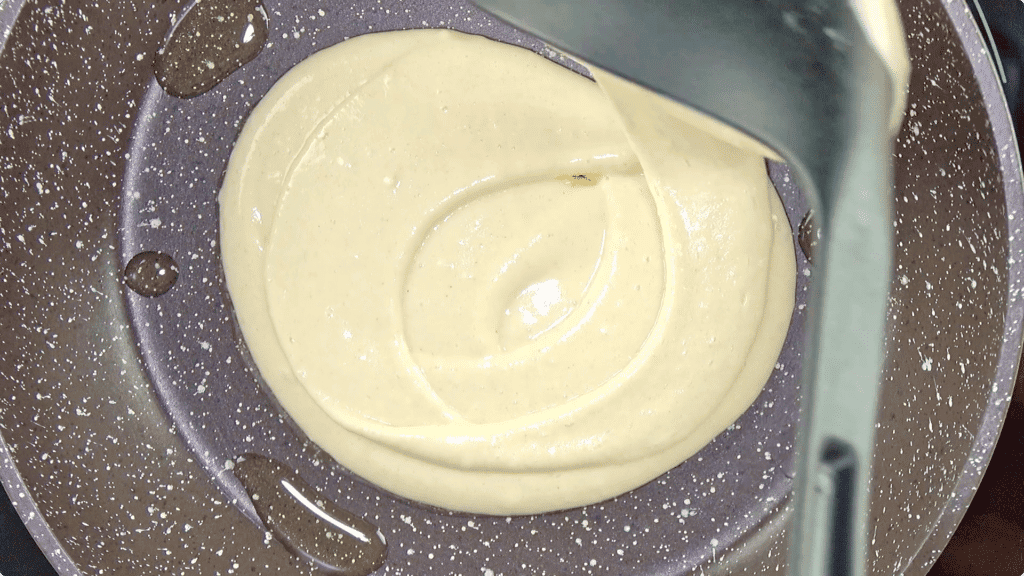 A spoonful of pancakes batter being cooked in a black pan with a splash of oil