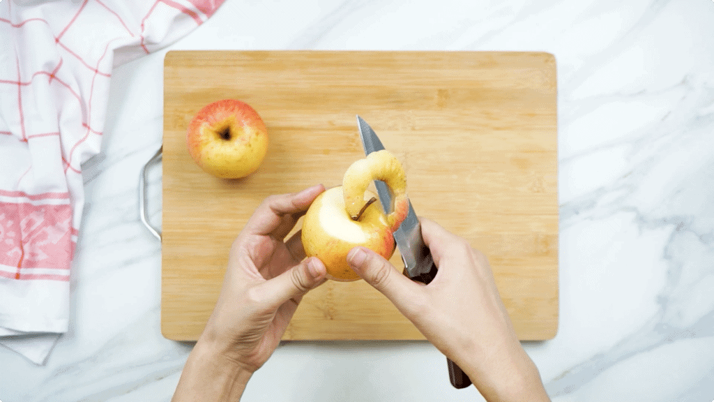 A woman's hands peeling an apple using a sharp knife with a wooden chopping board in the background