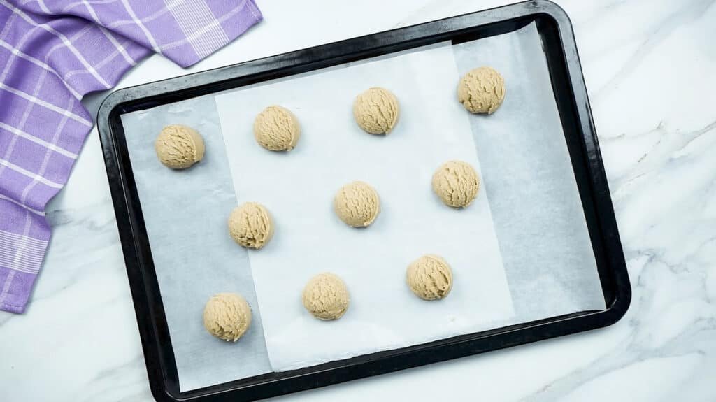 Cookie dough balls spaced out on a baking tray lined with parchment paper on a marble background