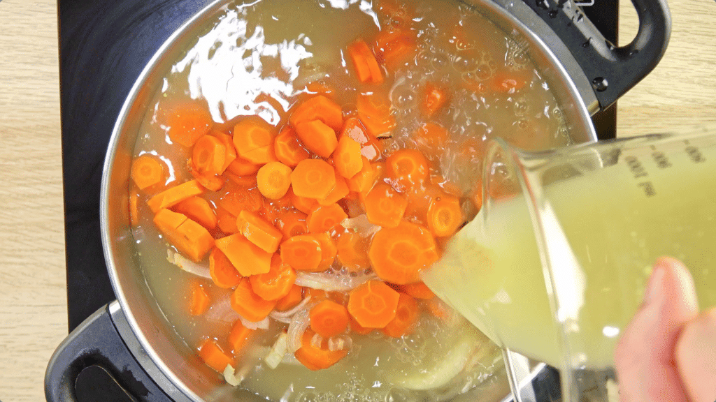 Carrot slices and onions in a pot with stock as more stock is added from a glass jar